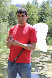 Dr. Rassim Khelifa stands in the foreground in a red t-shirt and jeans. He has a white insect net tucked under his arm, and is holding a dragonfly. He is in a wetland. 